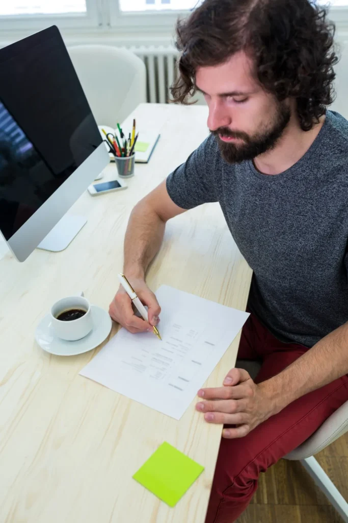 mężczyzna z brodą siedzi przy biurku i pisze coś na kartce przed monitorem komutera